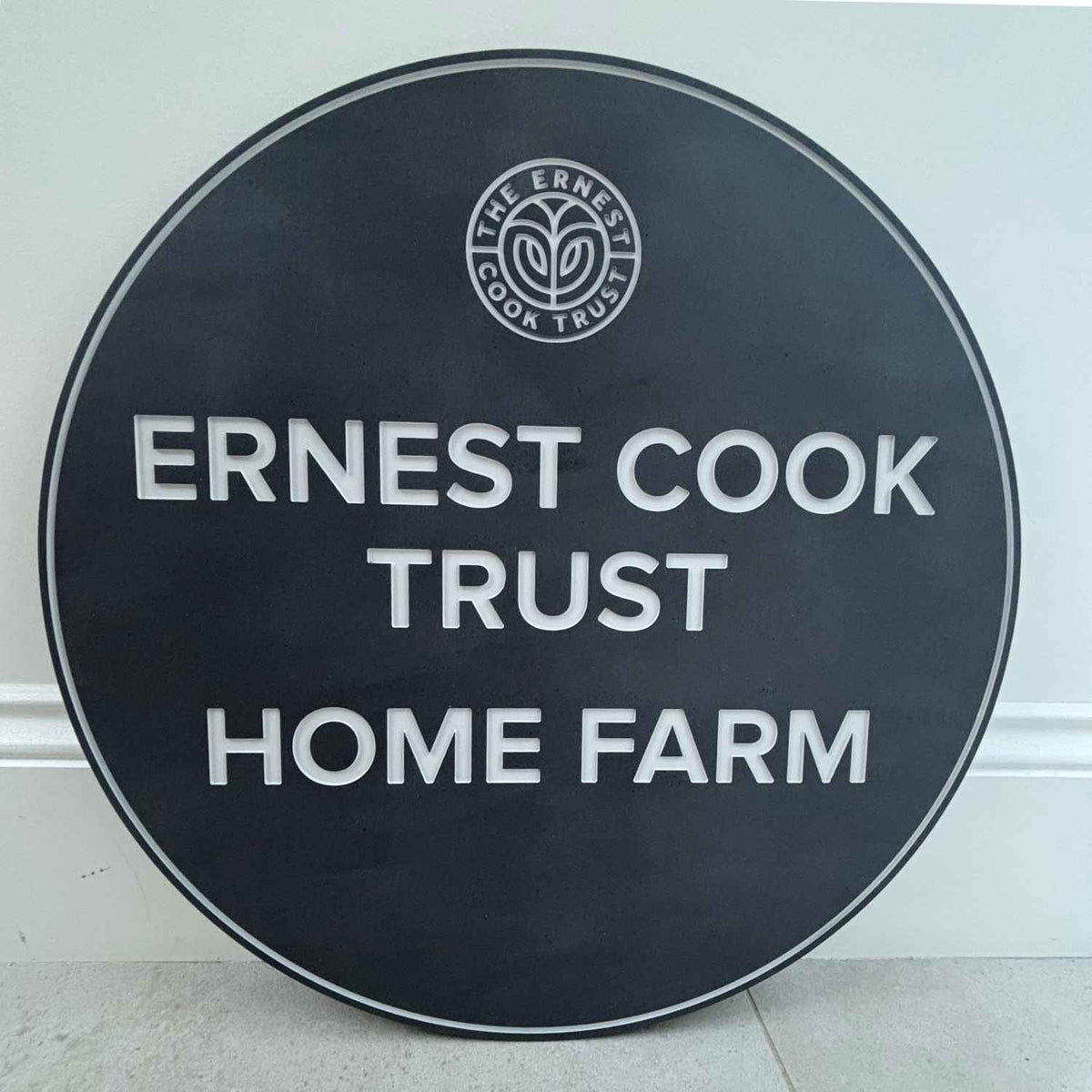 A round slate sign with white text reading 'ERNEST COOK TRUST HOME FARM' and a circular 'The Ernest Cook Trust' logo at the top, photographed leaning against a light wall.