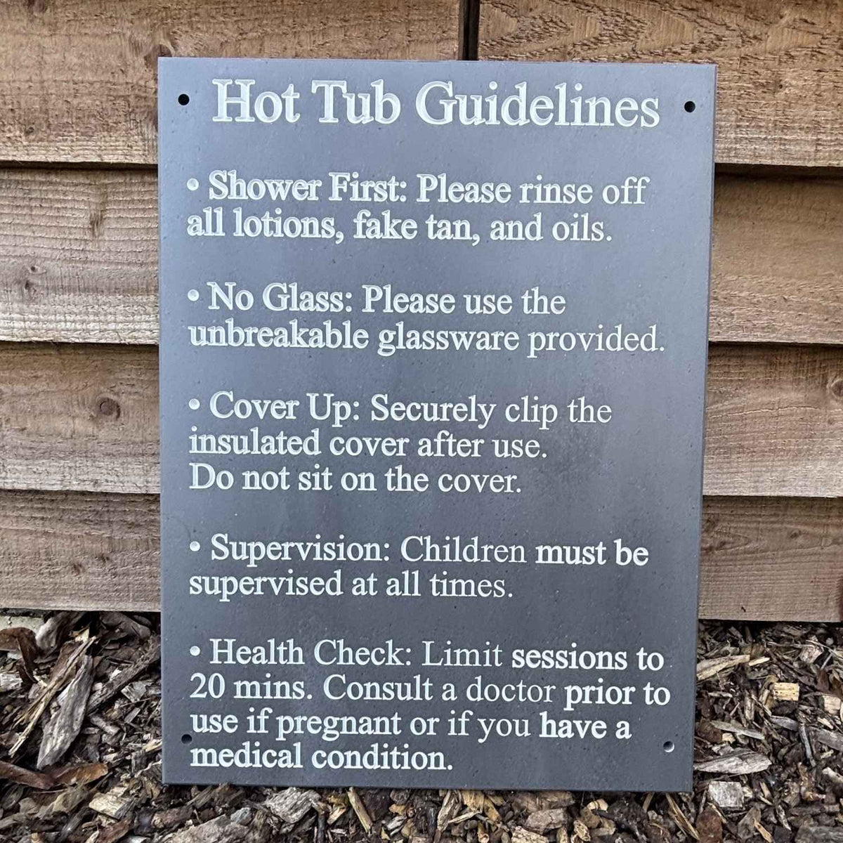 A rectangular slate sign with rounded corners reading 'Hot Tub Guidelines' in white text, mounted against a rustic wooden fence with mulch below. The sign lists safety instructions such as showering first, no glass, cover up, supervision, and health check.