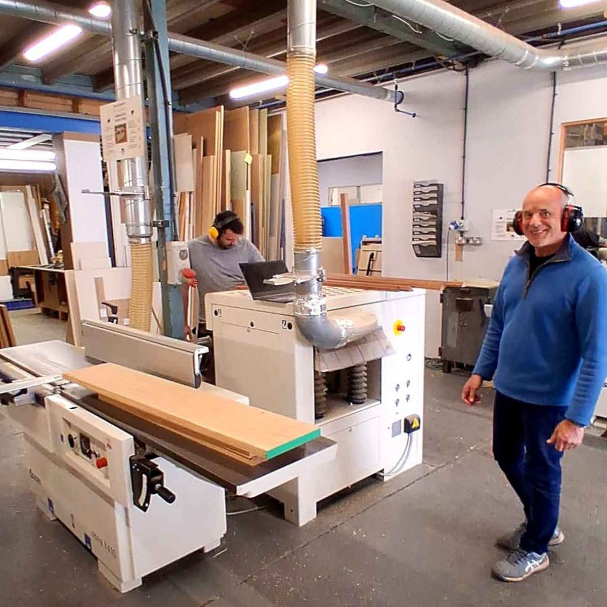 Man standing in a workshop with woodworking equipment and another person working in the background.