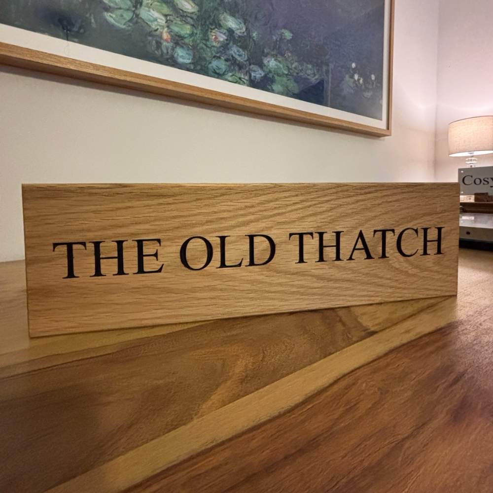 A horizontal oak ooden sign with the text 'THE OLD THATCH' in black serif lettering, displayed on a wooden table in an indoor room setting.