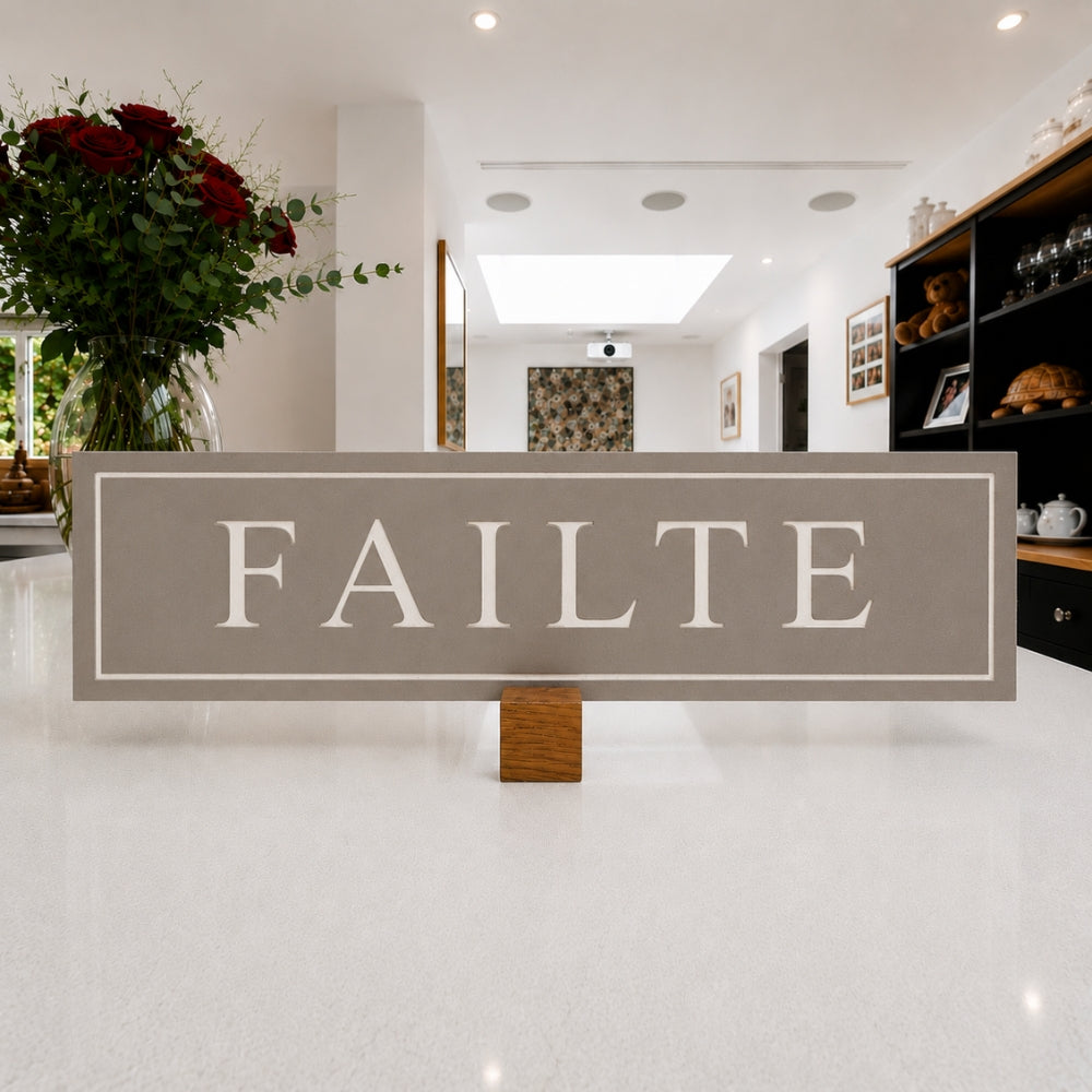 A rectangular grey sign with the word 'FAILTE' in large white serif letters, displayed on a small wooden block in a bright, modern interior hallway.
