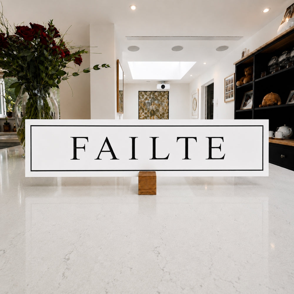 A rectangular white sign with the word 'FAILTE' in black serif letters, displayed on a small wooden stand in a bright, modern interior hallway with white marble flooring and decorative shelving.