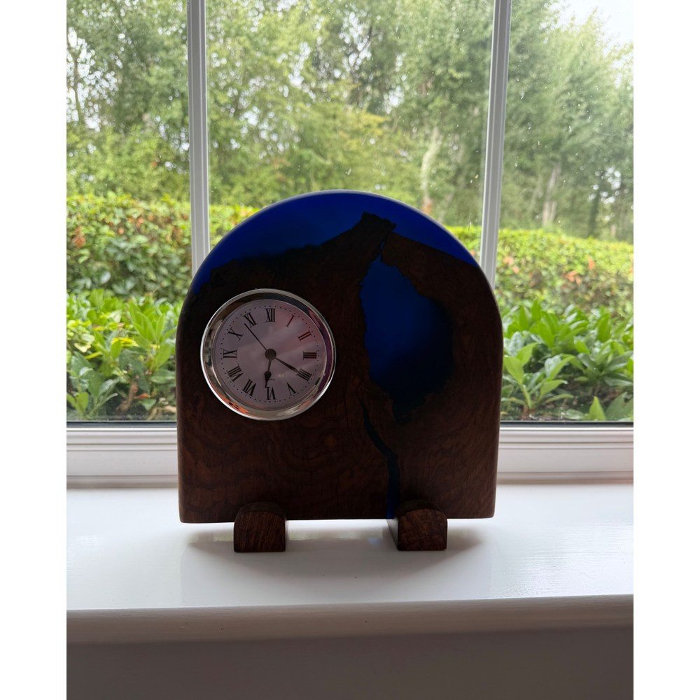 A small wooden mantel clock with a dark blue resin inlay, displayed on a white windowsill with a garden view in the background.
