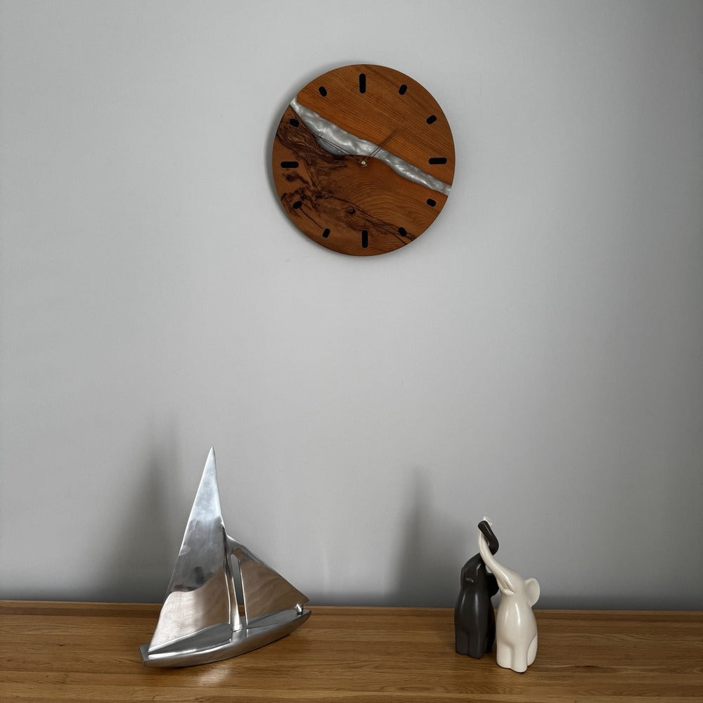 A round wooden wall clock with a split wood and silver metallic design, mounted on a light gray wall above a wooden surface with decorative objects.