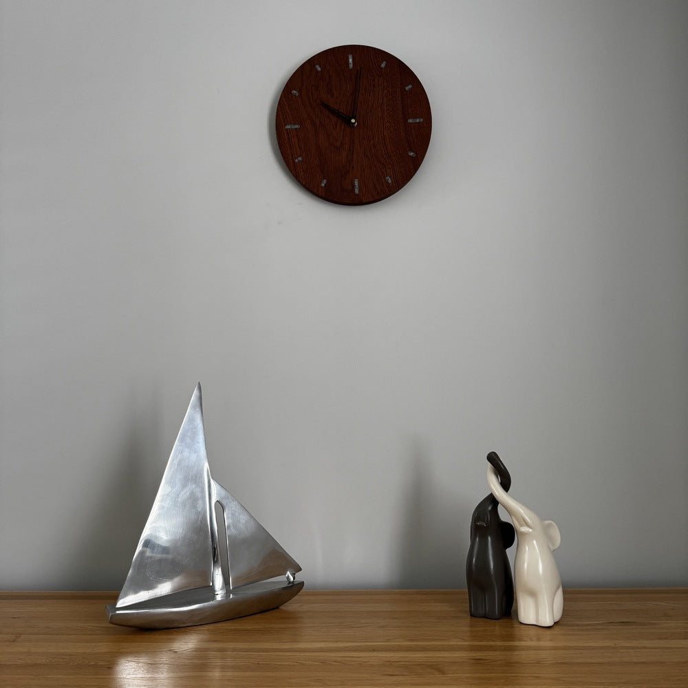 A round dark wood wall clock hangs on a light gray wall above a wooden table. On the table are a silver metallic sailboat sculpture and a pair of black and cream elephant figurines.