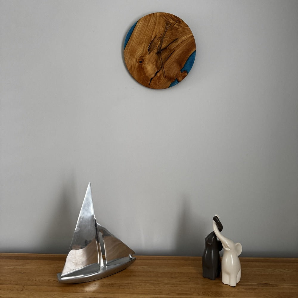 A round wooden wall clock with a natural live edge and blue resin accents, mounted on a light gray wall above a wooden console table with decorative sculptures.