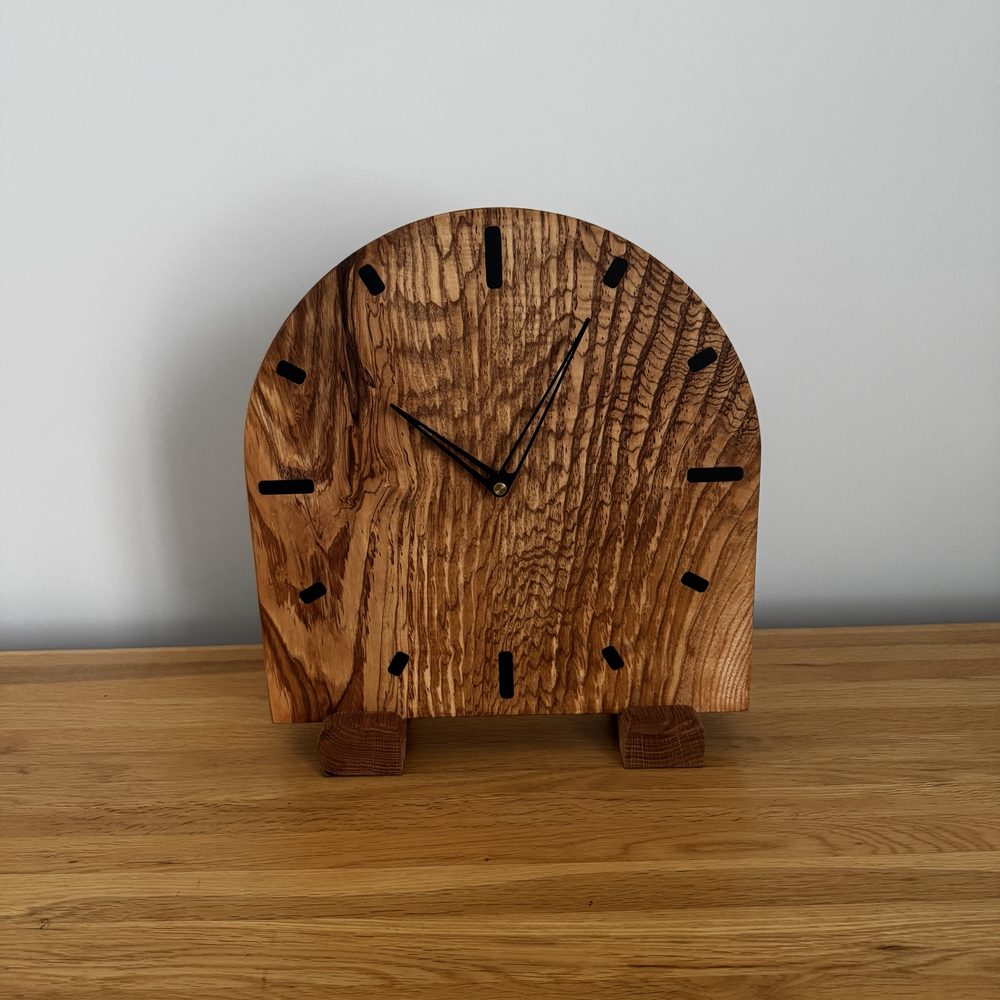 A rustic wooden tabletop clock with a rounded arch shape, featuring prominent natural wood grain, black hour markers, and black hands, displayed on a wooden surface against a light gray wall.