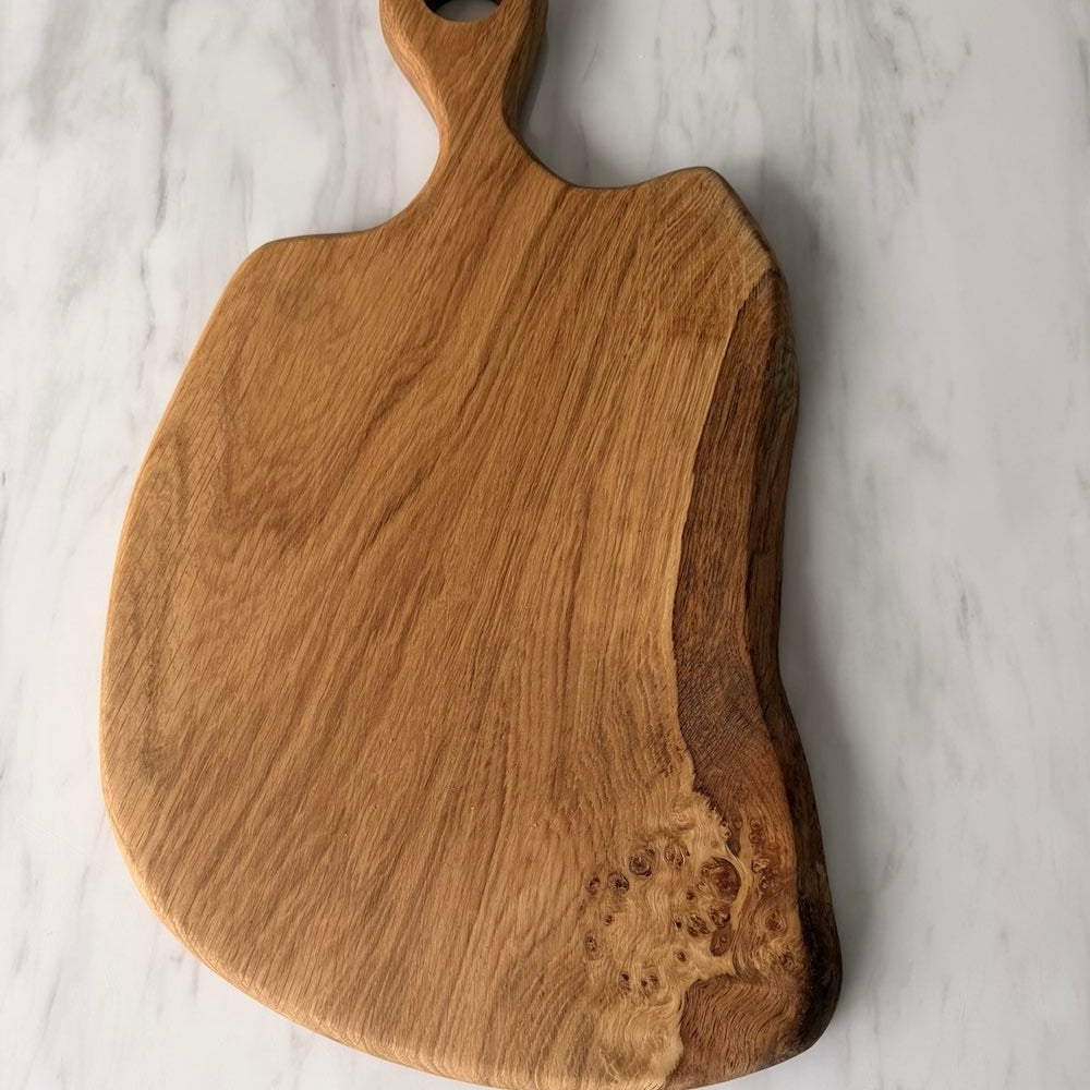 A rustic handcrafted wooden cutting board with a natural live edge and a handle, displayed on a white marble surface.