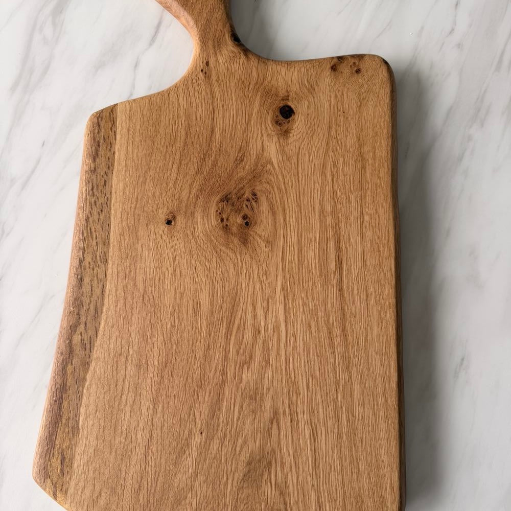 A natural wood cutting board with a handle, displayed diagonally on a white marble surface.