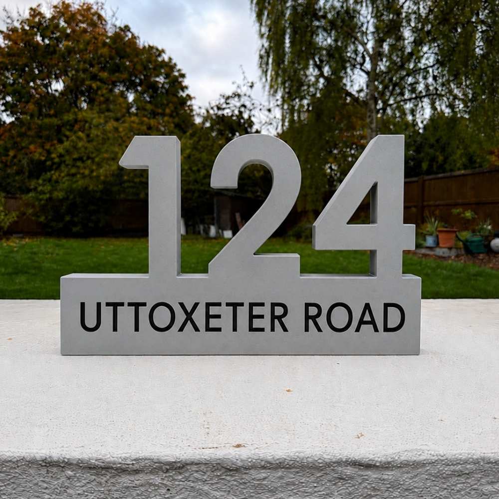 A modern house number sign displaying '124 UTTOXETER ROAD' in black lettering on a brushed silver metal base, set outdoors on a concrete ledge with a garden background.