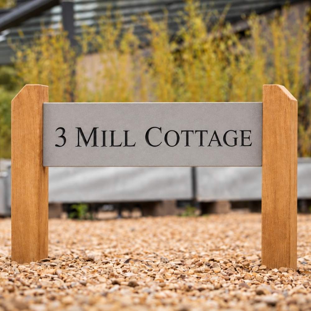 A custom engraved stone sign reading '3 MILL COTTAGE' mounted on a wooden frame, set in gravel with a blurred garden background.