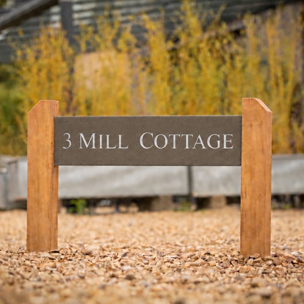 A rectangular mink sign with the words “3 MILL COTTAGE” in white lettering, mounted between two natural wood posts on a gravel surface outdoors.