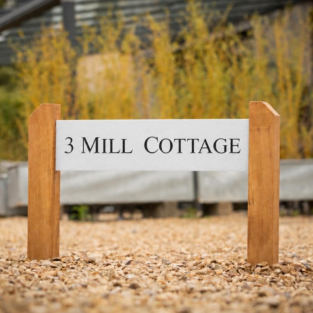 A rectangular white sign with black text reading '3 MILL COTTAGE' mounted between two wooden posts, set outdoors on a gravel surface with blurred plants in the background.