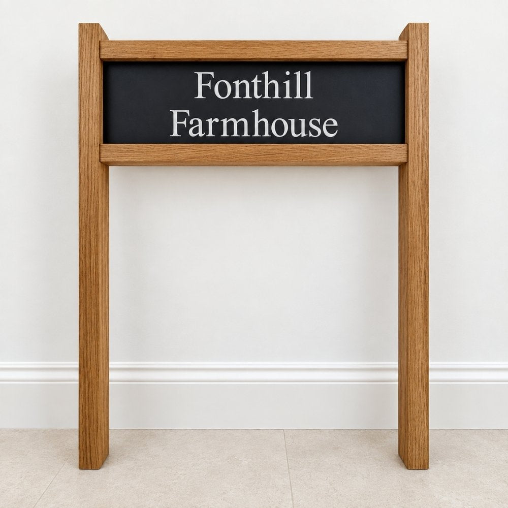 A wooden freestanding sign with a dark slate rectangular panel reading 'Fonthill Farmhouse' in white serif text, shown against a plain light wall and pale floor.
