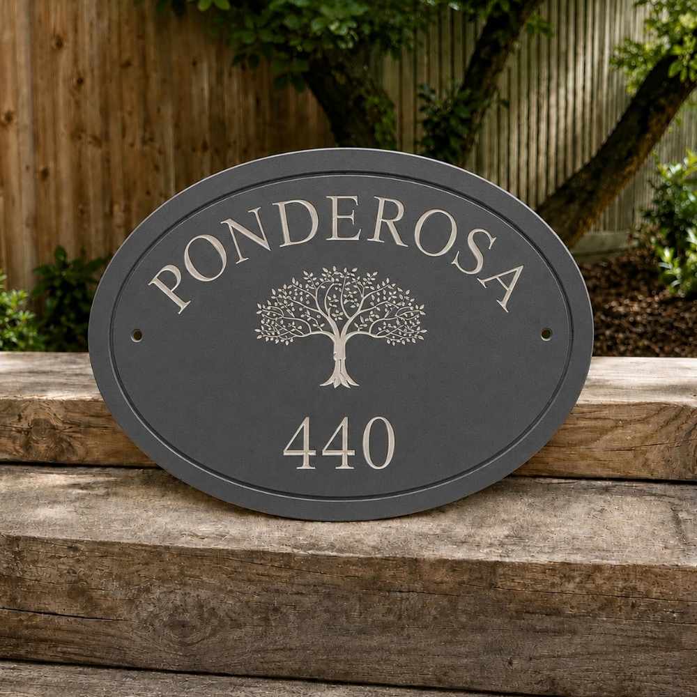 An oval slate house plaque with the name 'PONDEROSA' and number '440' in silver, featuring a tree design in the center, mounted on a wooden fence in a garden setting.