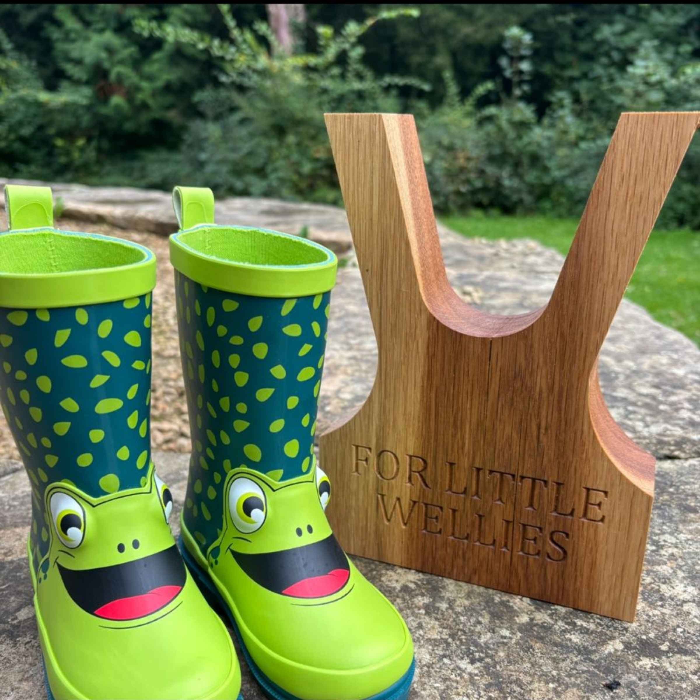 Green children's wellies with frog design next to a wooden boot jack engraved 'FOR LITTLE WELLIES'.