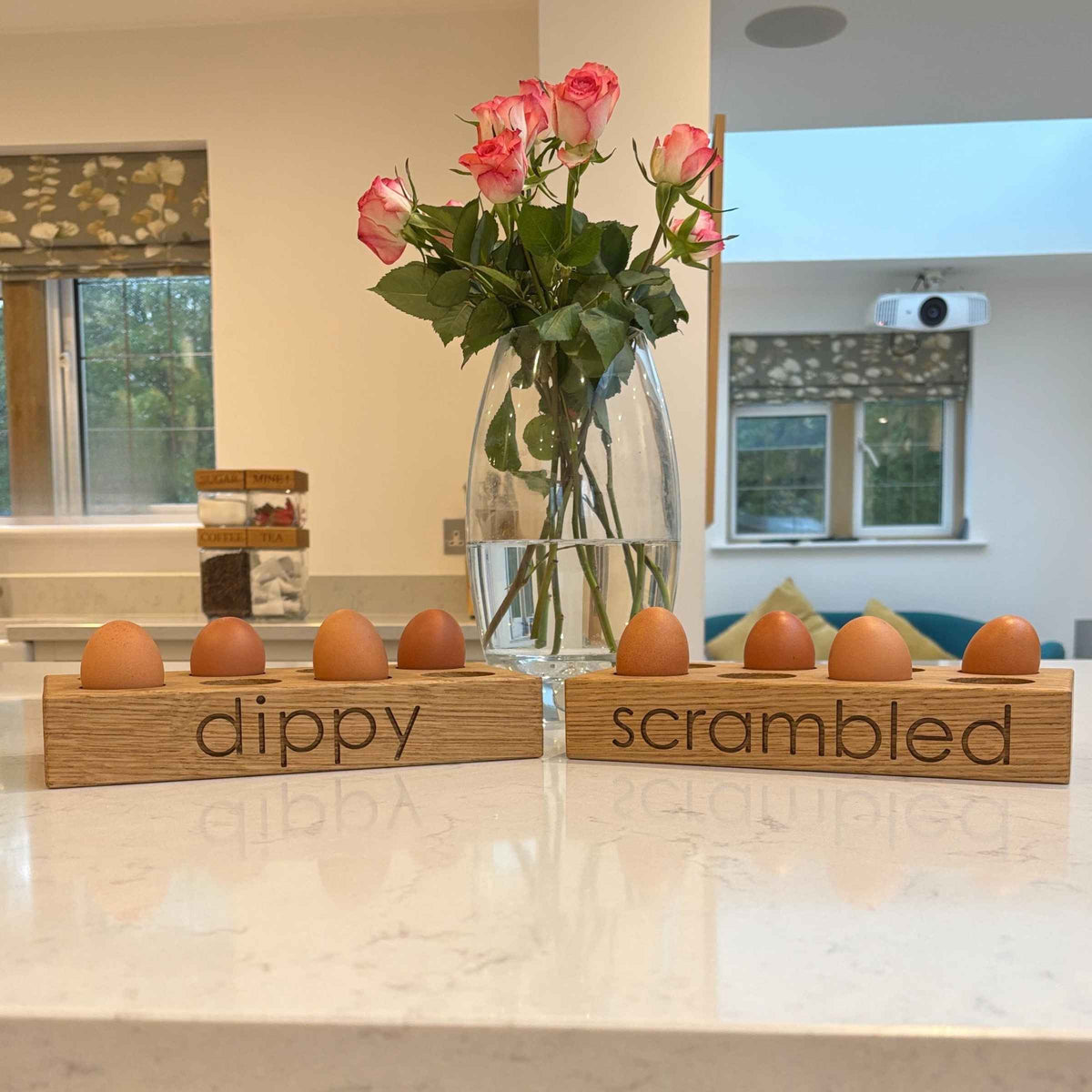 Two wooden egg holders labeled 'dippy' and 'scrambled' with a vase of pink roses in the background.