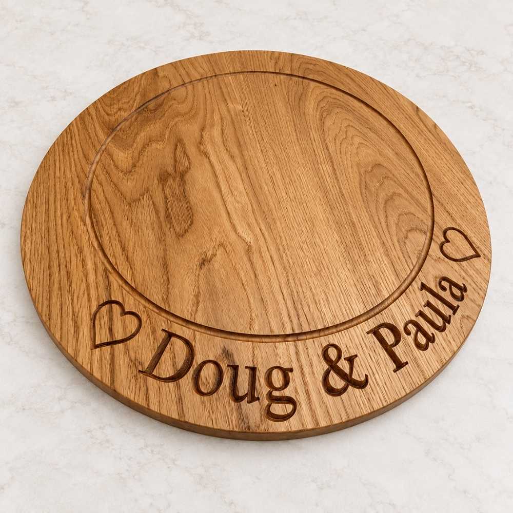 A round wooden serving board with engraved names and hearts, shown on a light marble surface. The board is personalized with the text 'Doug & Paula' and two heart icons.