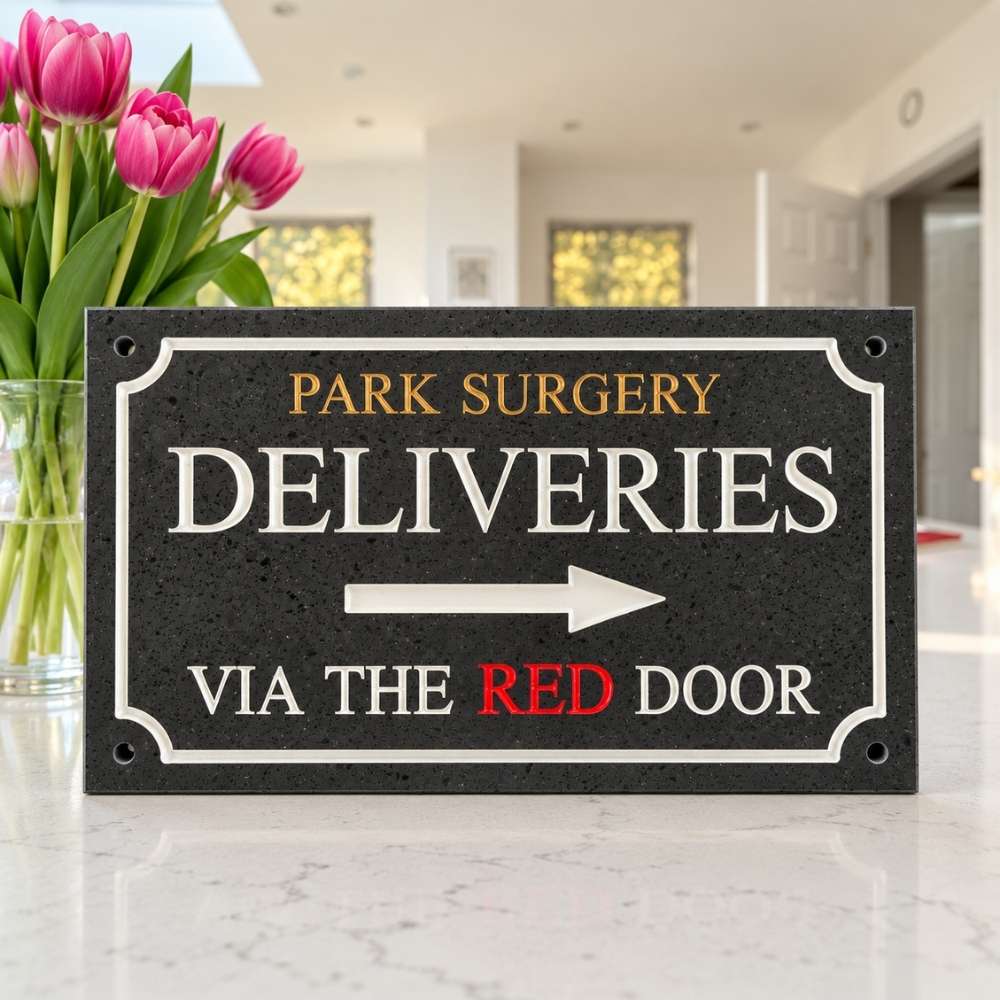A rectangular charcoal stone delivery sign for Park Surgery, mounted on a white marble countertop. The sign features white and gold lettering that reads 'PARK SURGERY DELIVERIES' with a white arrow pointing right, and 'VIA THE RED DOOR' in white and red.