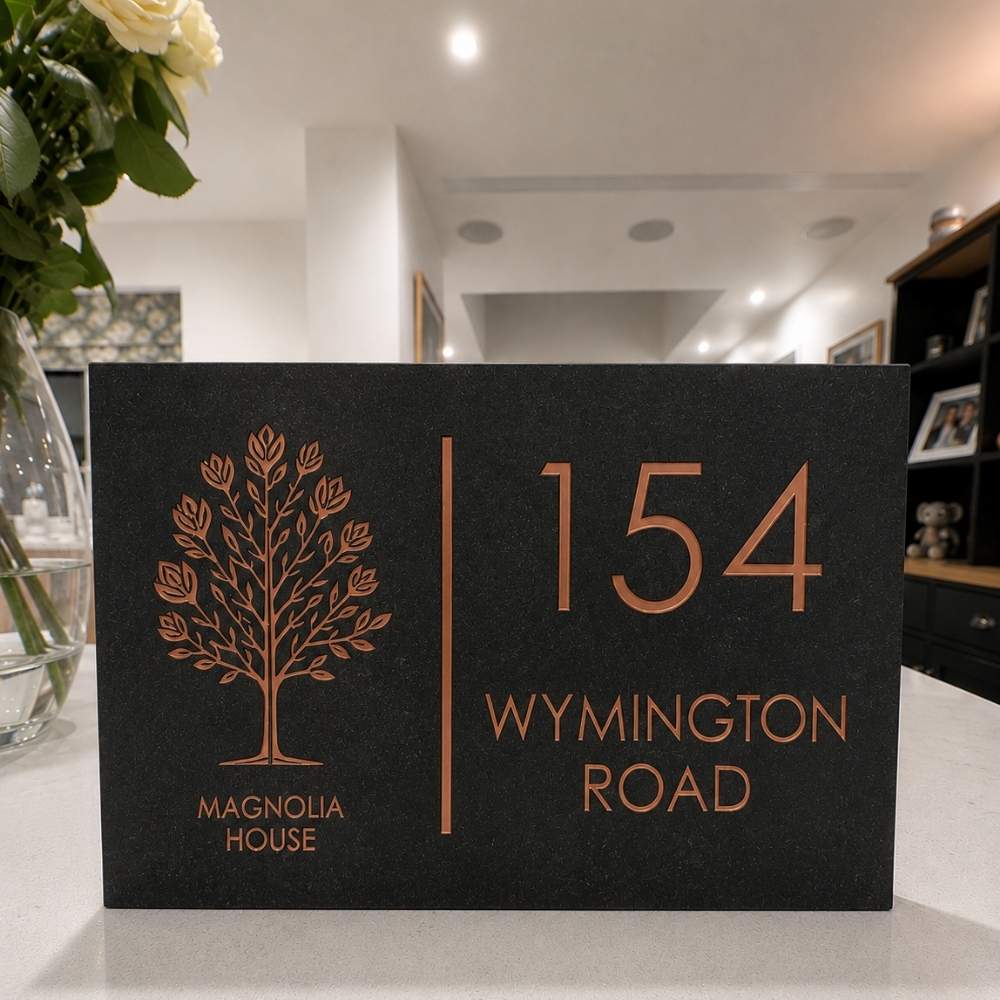 A charcoal house number sign with rose gold lettering and a tree emblem, reading '154 WYMINGTON ROAD' and 'MAGNOLIA HOUSE', displayed on a countertop in a home interior.