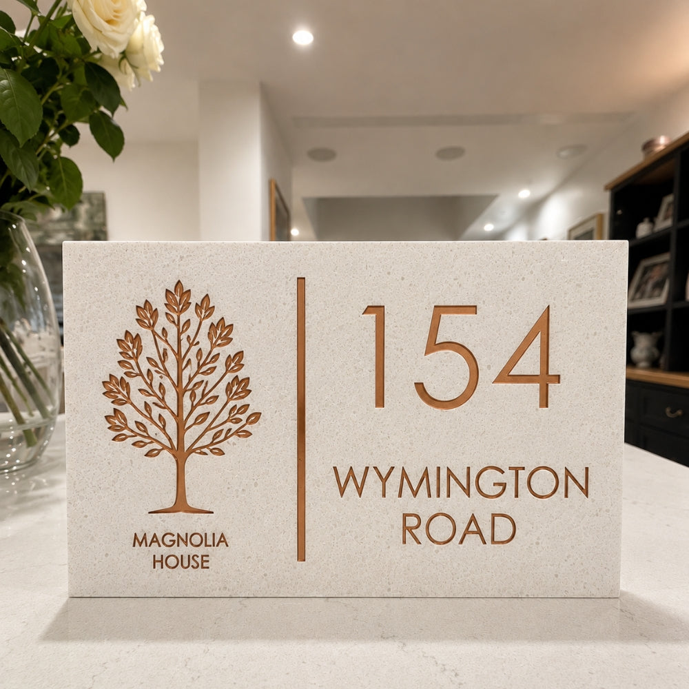 A rectangular limestone stone house number plaque with a bronze tree logo and the text 'MAGNOLIA HOUSE' and '154 WYMINGTON ROAD', displayed on a countertop in a home interior.
