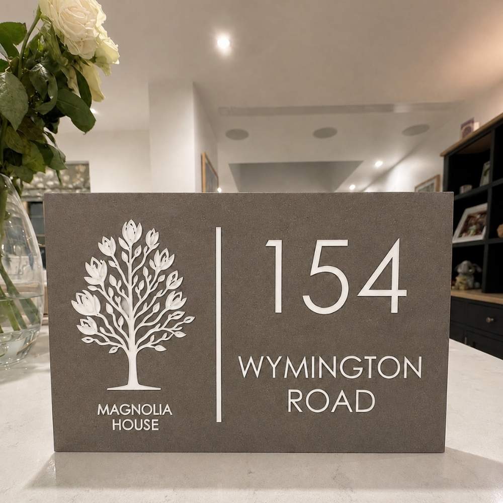 A rectangular mink house number sign with a white tree logo and the text 'MAGNOLIA HOUSE' and '154 WYMINGTON ROAD', displayed on a light countertop in a home interior.