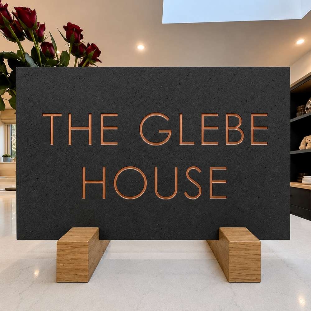 A rectangular charcoal stone sign with engraved copper-toned lettering reading 'THE GLEBE HOUSE,' displayed on two light wood blocks in an indoor setting with flowers and shelves in the background.