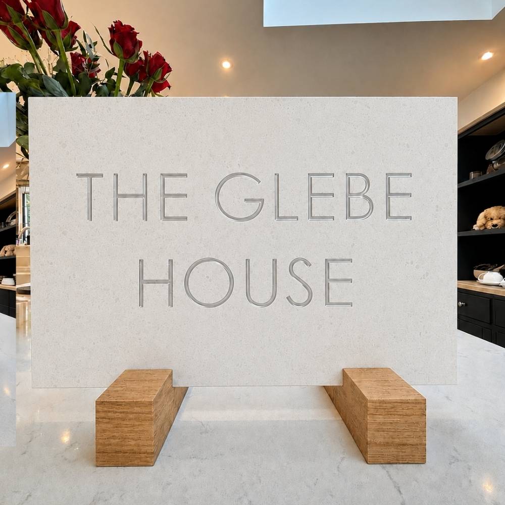 A quartz rectangular cream stone or concrete sign with the words “THE GLEBE HOUSE” in raised gray lettering, displayed on two wooden blocks on a light marble surface in an indoor setting.