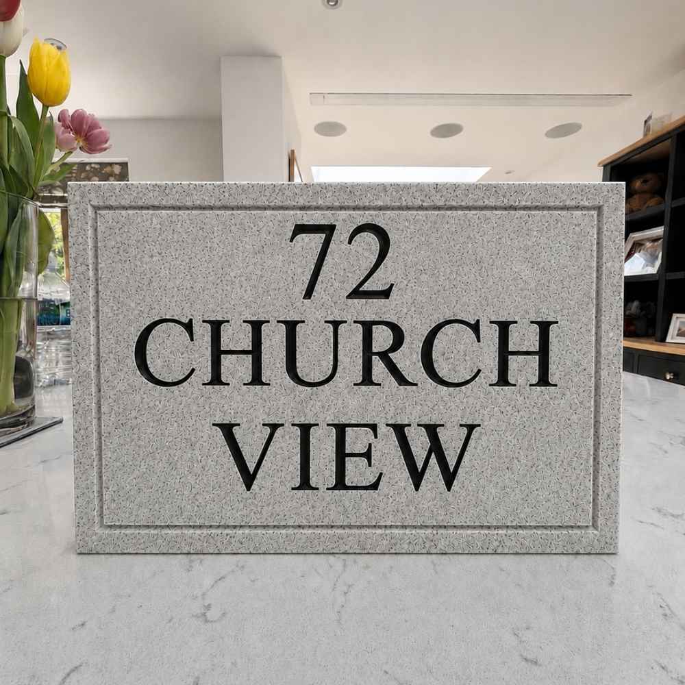 A rectangular granite stone or concrete plaque with the text '72 CHURCH VIEW' in black serif lettering, placed on a light marble countertop in a real indoor setting with flowers and shelving visible in the background.