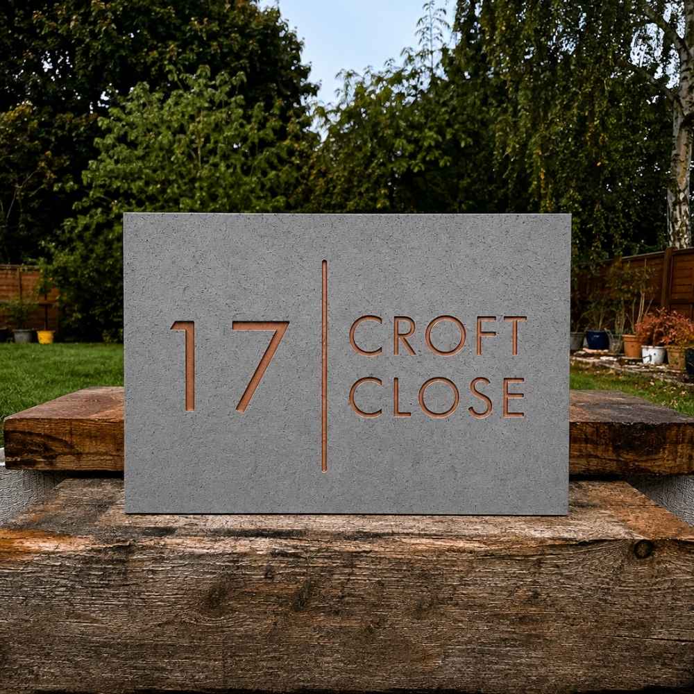 A rectangular granite stone house number plaque reading '17 CROFT CLOSE' with a vertical divider and engraved copper-toned lettering, displayed outdoors on a rustic wooden post with trees and a garden in the background.