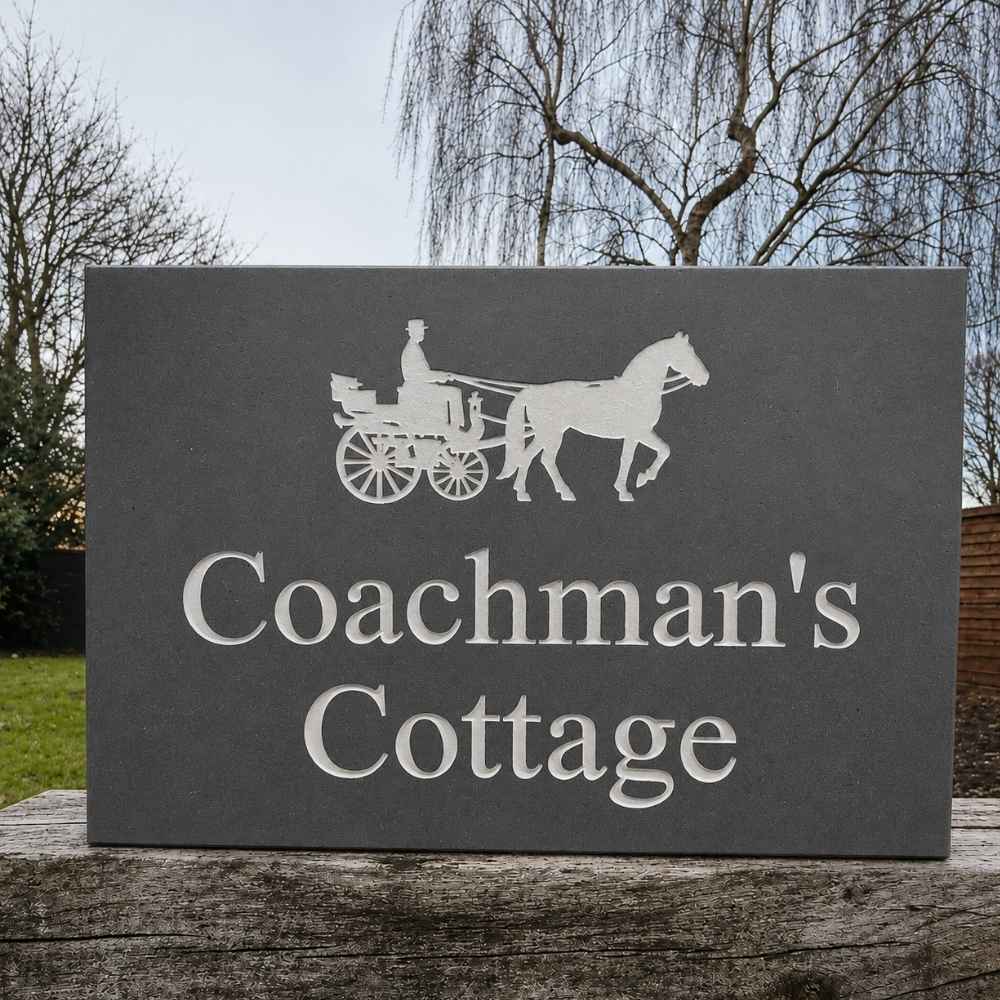 A rectangular slate sign with a white silhouette of a horse-drawn carriage and the words 'Coachman's Cottage' in large white lettering, photographed outdoors on a wooden surface with trees and grass in the background.
