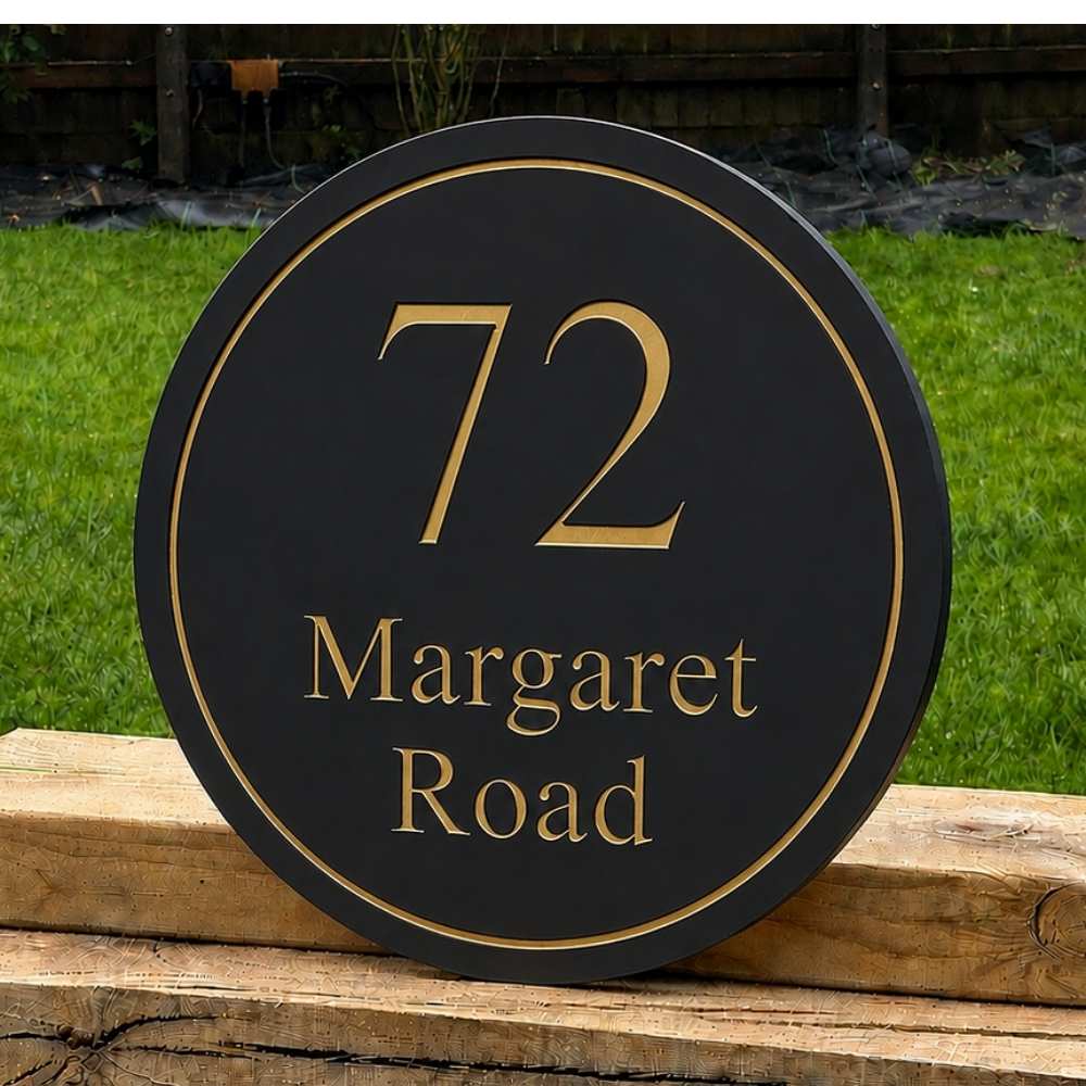 A round black house number sign with gold lettering reading '72 Margaret Road' displayed outdoors on a wooden fence with green grass in the background.