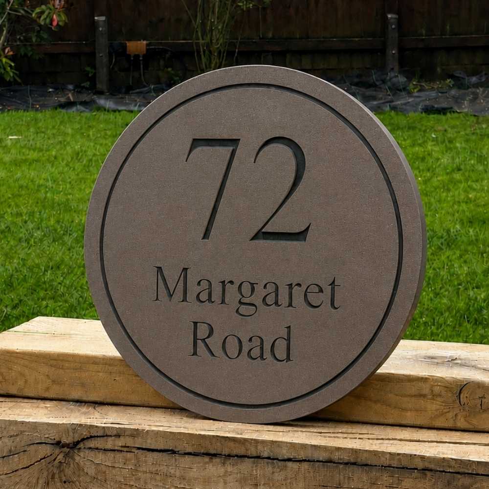 A round mink house number plaque reading '72 Margaret Road' in black lettering, displayed outdoors on a wooden fence with green grass in the background.