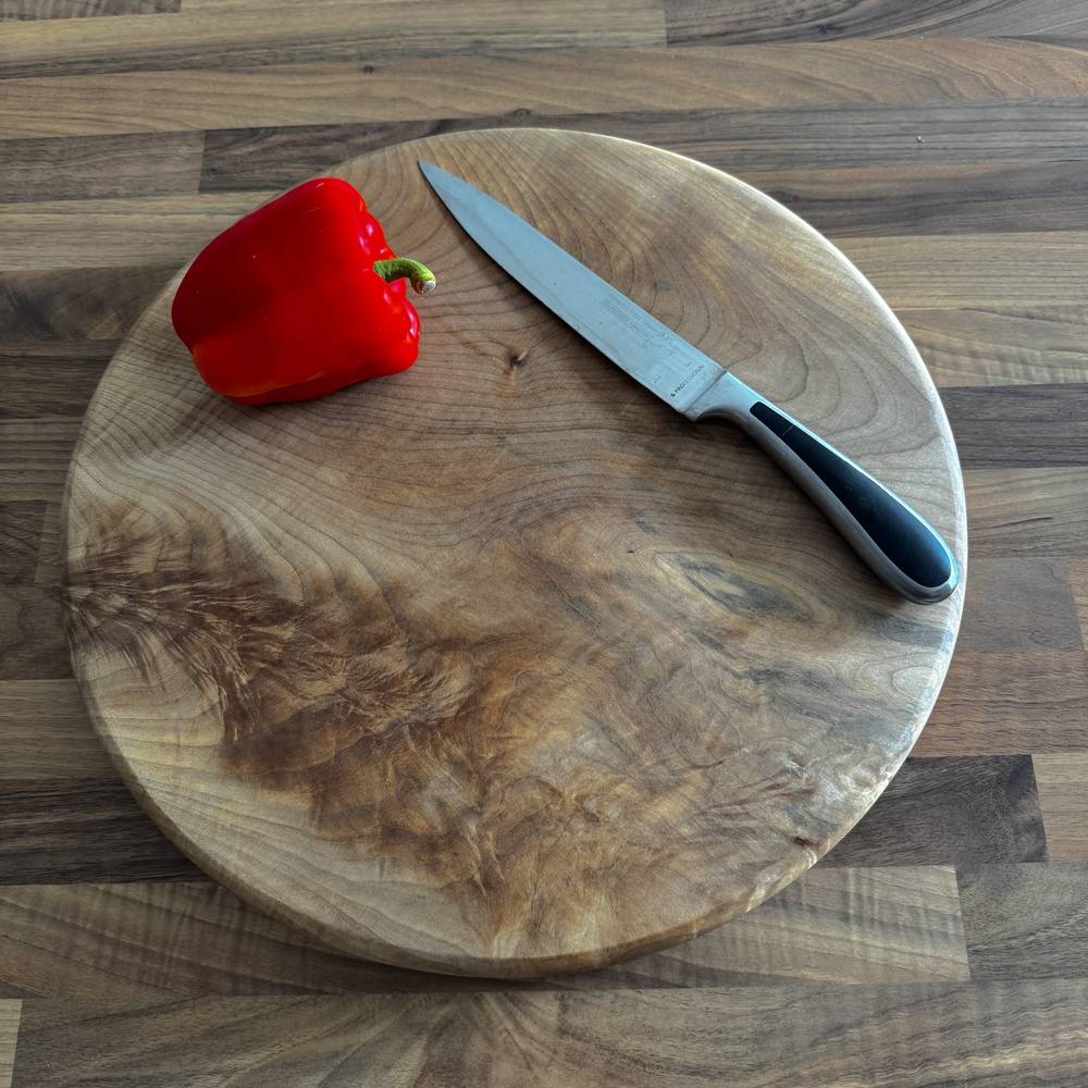 A round wooden cutting board with a natural grain pattern, shown on a matching wood countertop. A red bell pepper and a chef's knife with a black handle rest on the board.
