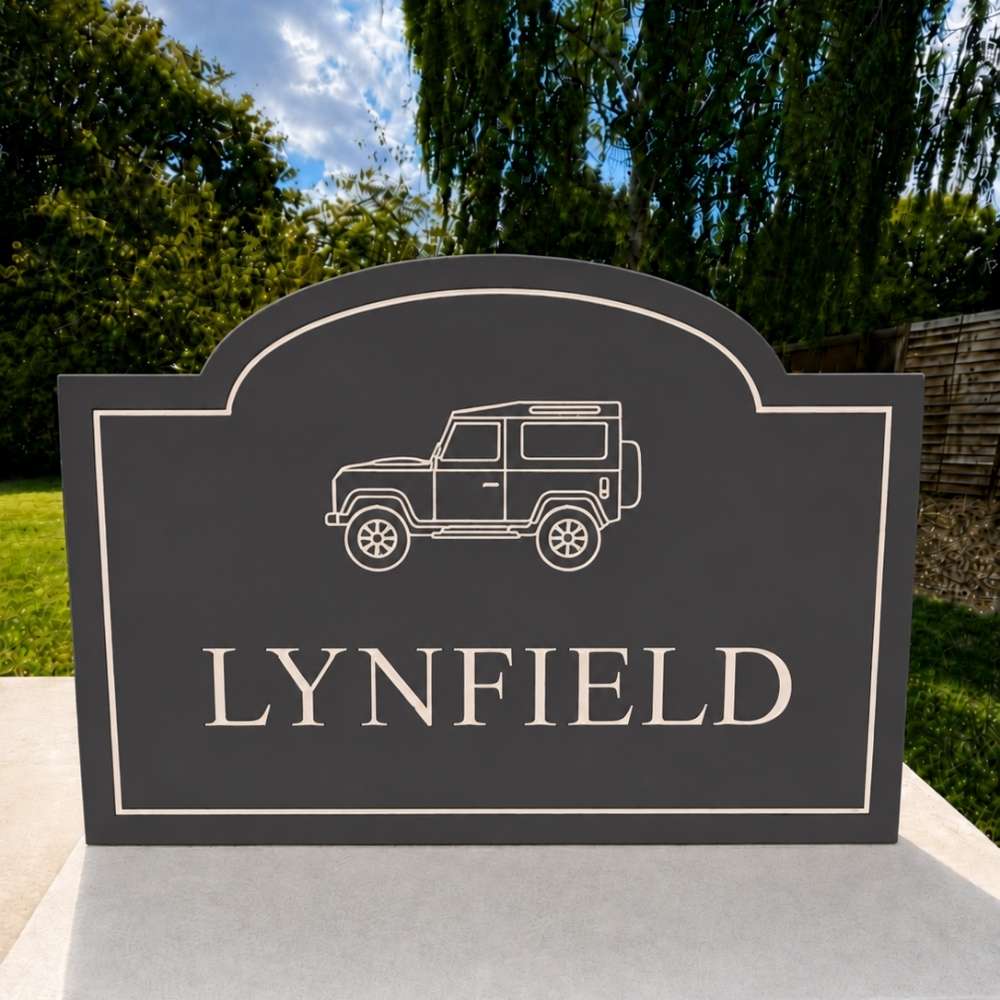A dark slate sign with an engraved white outline of a classic off-road vehicle and the name 'LYNFIELD' in large capital letters, displayed outdoors on a light-colored stone pedestal with a garden background.