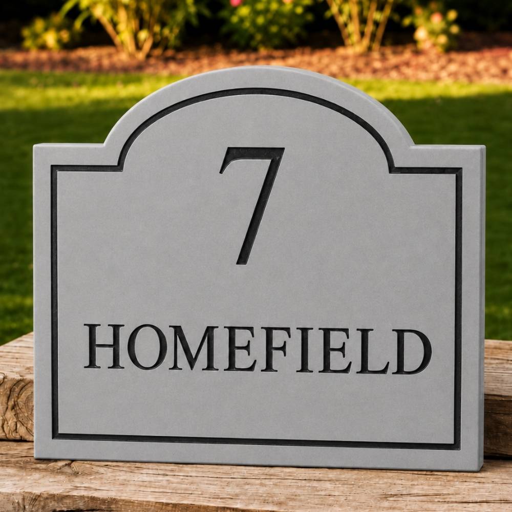 A grey arched house number plaque with the number 7 and the word HOMEFIELD in black, displayed on a wooden post outdoors with a blurred green lawn and garden background.