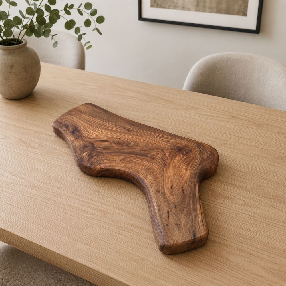 A rustic live-edge wooden serving board with a natural irregular shape and rich dark brown grain, displayed on a light oak dining table in a modern, neutral-toned room.