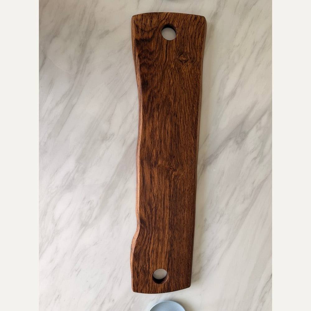 A long, narrow wooden serving board or cutting board with a rich dark brown wood grain, featuring two circular cutouts near the top and bottom, displayed on a white marble surface.