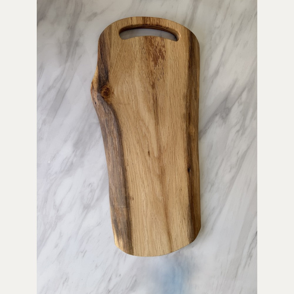 A natural live-edge wooden cutting board with a handle cutout, displayed on a white and gray marble surface.