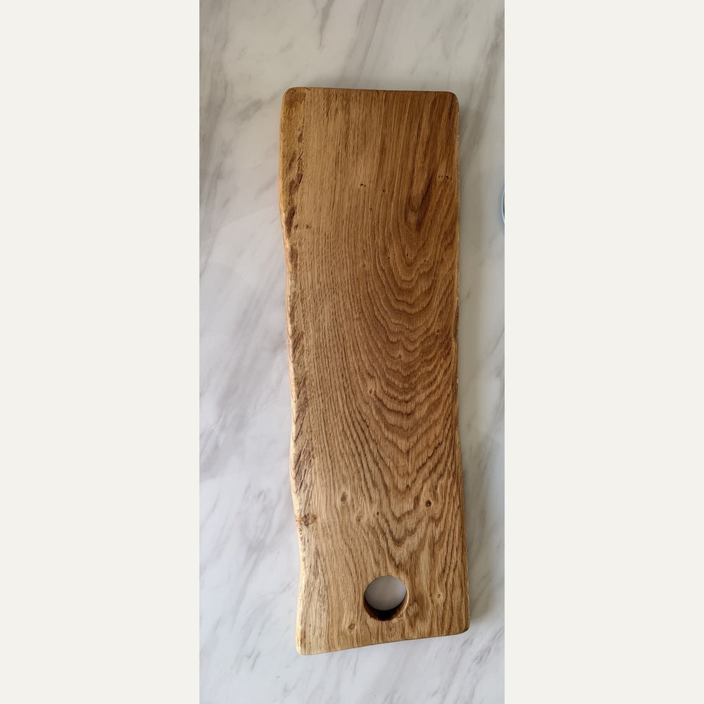 A rectangular wooden serving board with a live edge and a circular hole near one end, shown on a light marble surface.