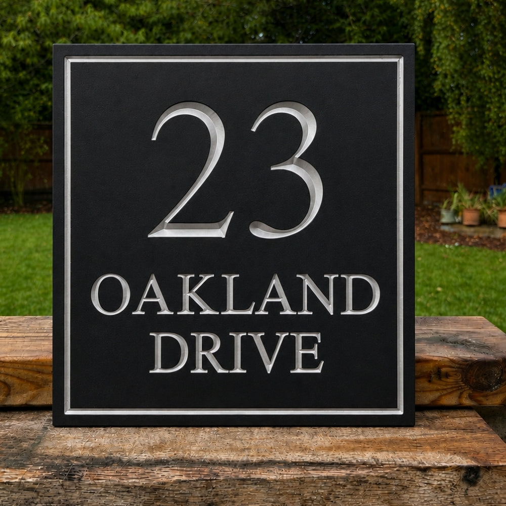 A square black house number sign with raised silver lettering reading '23 OAKLAND DRIVE', displayed outdoors on a wooden surface with green grass and trees in the background.