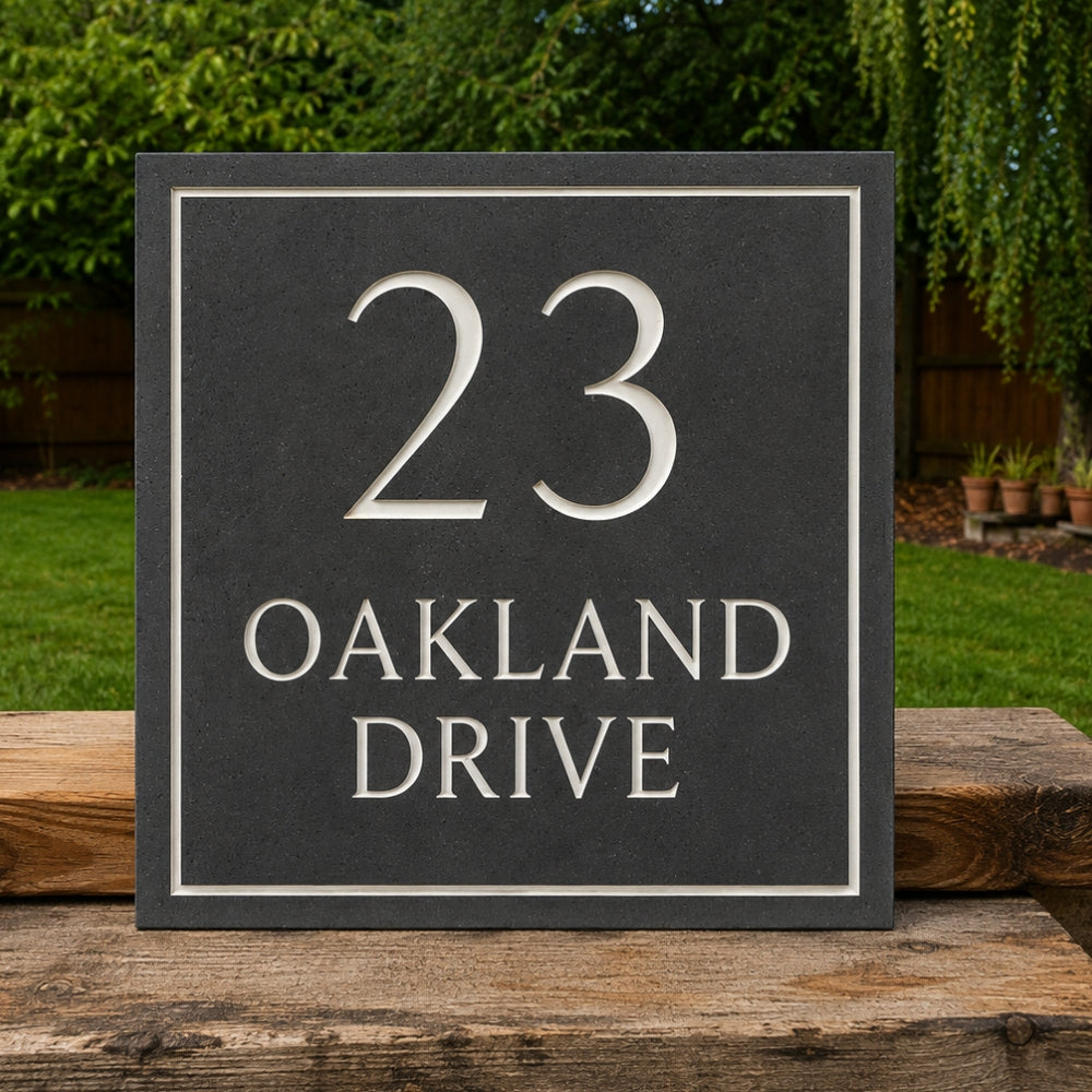 A square slate gray house number plaque reading '23 OAKLAND DRIVE' in raised silver lettering, displayed outdoors on a wooden surface with green grass and trees in the background.
