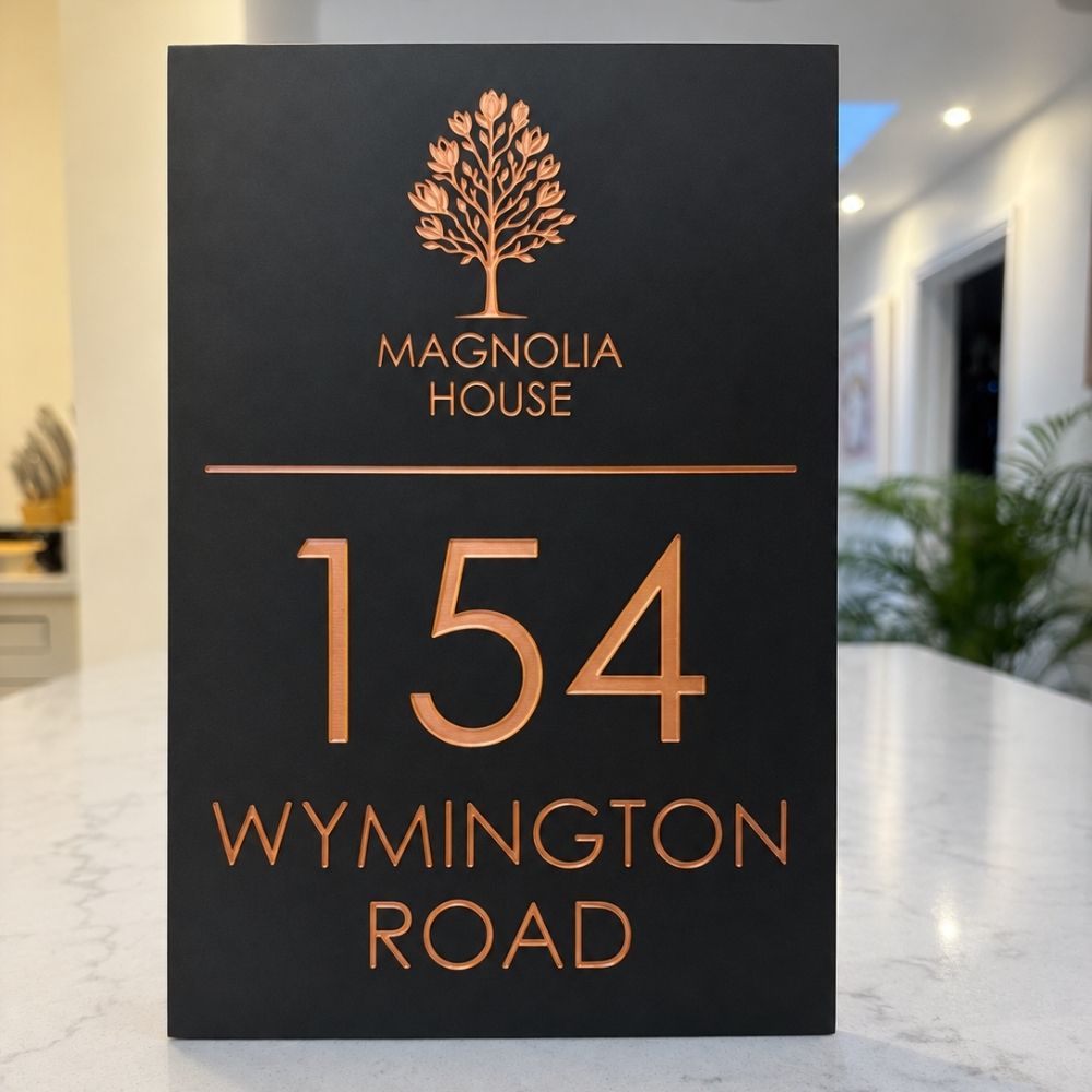 A black house number sign for Magnolia House at 154 Wynington Road, featuring a rose gold tree logo and lettering, displayed on a light marble countertop with a blurred interior background.
