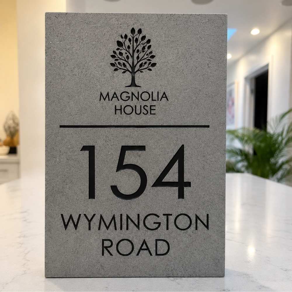 A rectangular charcoal stone house sign with black engraved text reading 'MAGNOLIA HOUSE', '154', and 'WYMINGTON ROAD', standing on a light countertop in a blurred indoor setting.