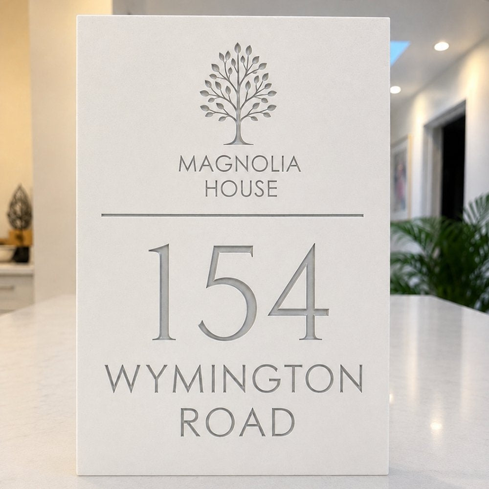 A white rectangular house sign with engraved gray text reading 'MAGNOLIA HOUSE', '154', and 'WYMINGTON ROAD', standing on a light countertop in a blurred indoor setting.