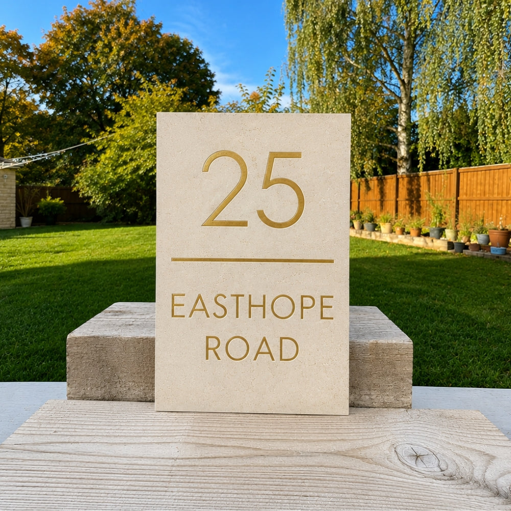 A rectangular limestone house number plaque reading '25 EASTHOPE ROAD' in gold lettering, displayed outdoors on a wooden surface with a green lawn and trees in the background.