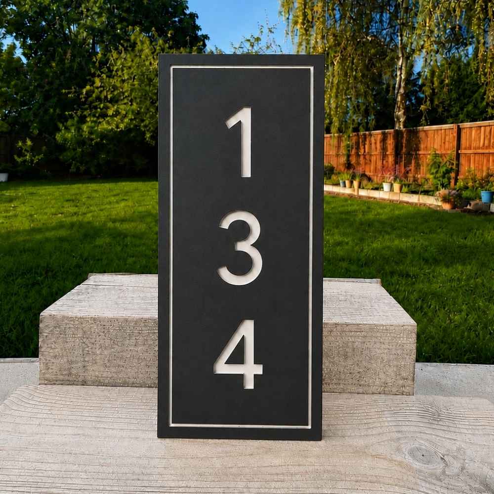 A vertical dark slate house number sign with white numerals '134' and a thin white border, shown outdoors on a stone riser with a green lawn and trees in the background.