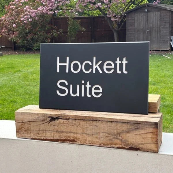 Sign reading 'Hockett Suite' on a wooden base outdoors with grass and a shed in the background.
