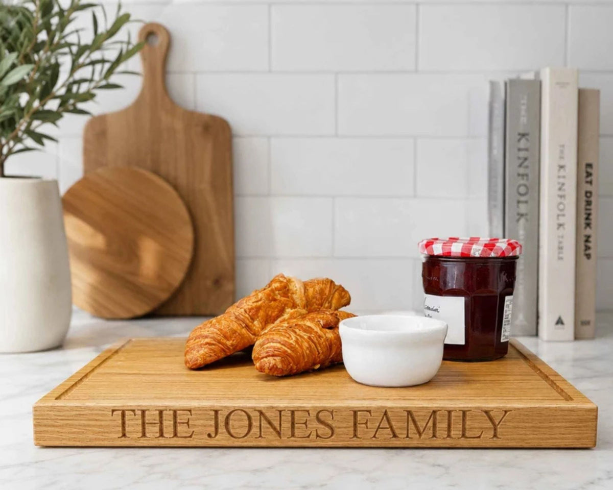 A rectangular wooden chopping board engraved with 'THE JONES FAMILY' sits on a marble countertop in a bright kitchen. On the board are two croissants and a small white bowl, with a jar of jam and cutting boards in the background.