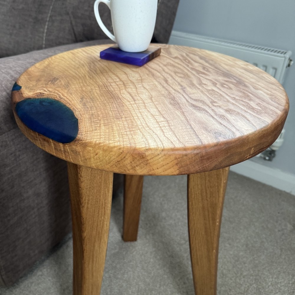 A round wooden end table with a natural live edge and dark blue resin inlay, shown in a living room setting next to a gray sofa with a white mug on top.